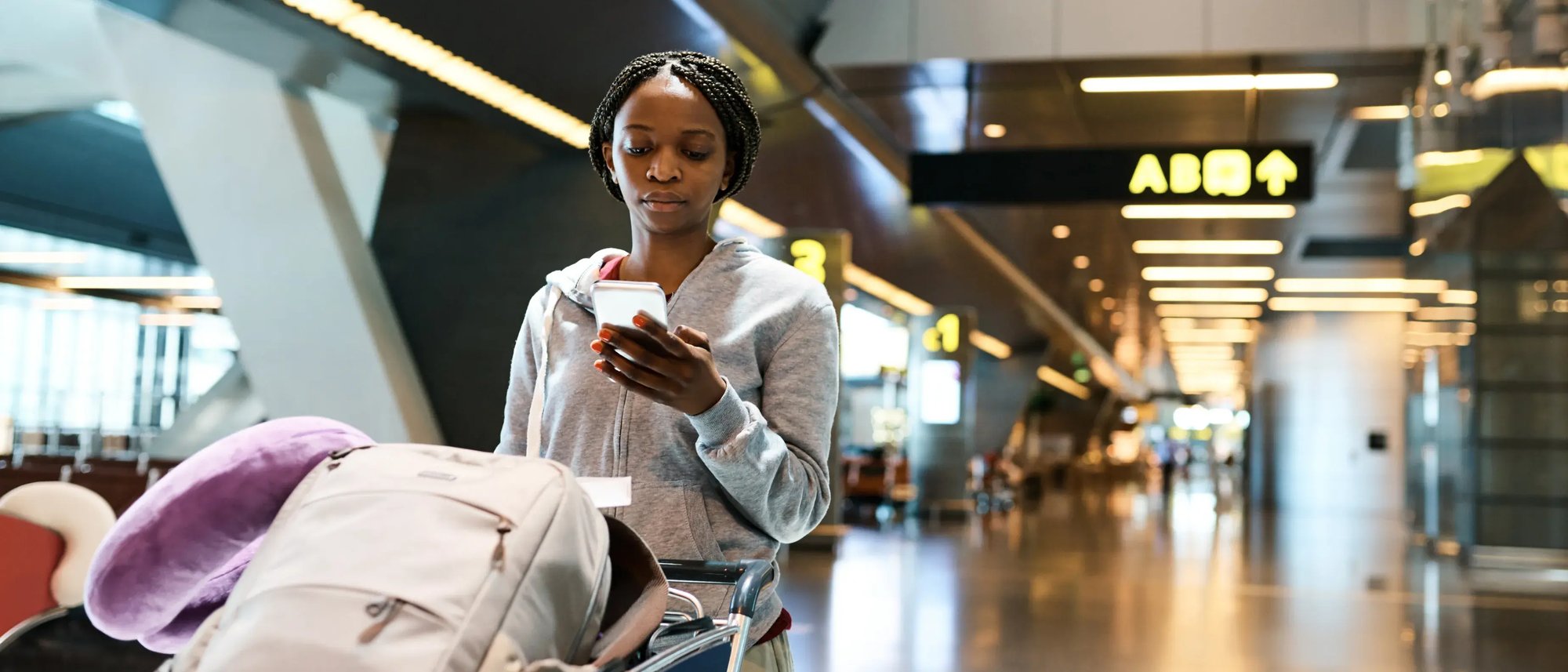 A woman looking down at her phone while pushing a cart at an airport.